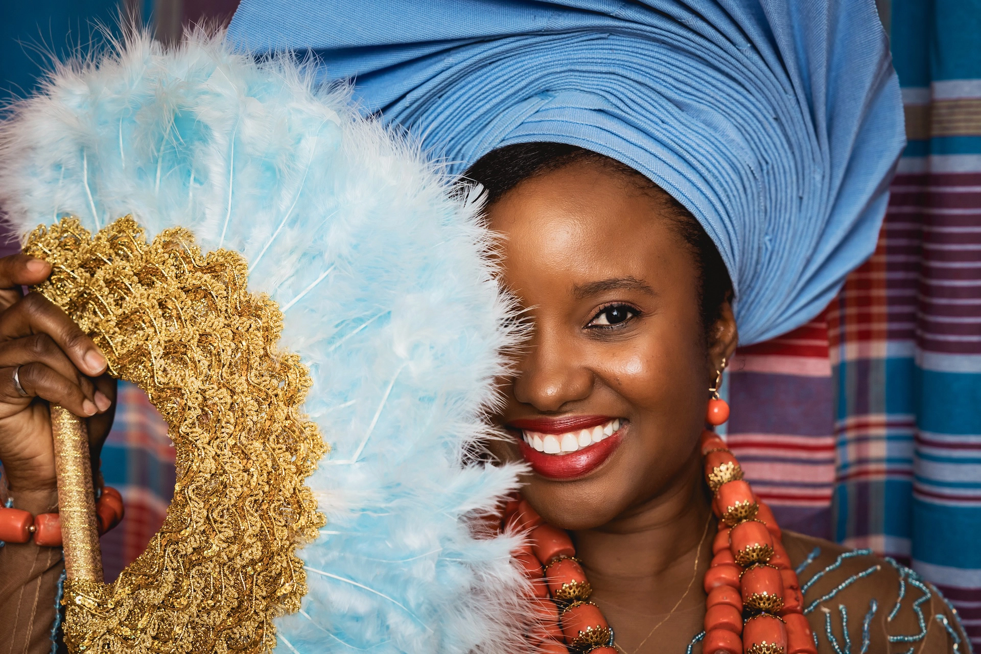 A stunning close-up of a bride in traditional African attire, captured during a vibrant wedding ceremony in Hamilton. Her radiant smile and half-hidden face behind a decorative fan bring elegance and emotion to this Southern Ontario wedding photography moment.