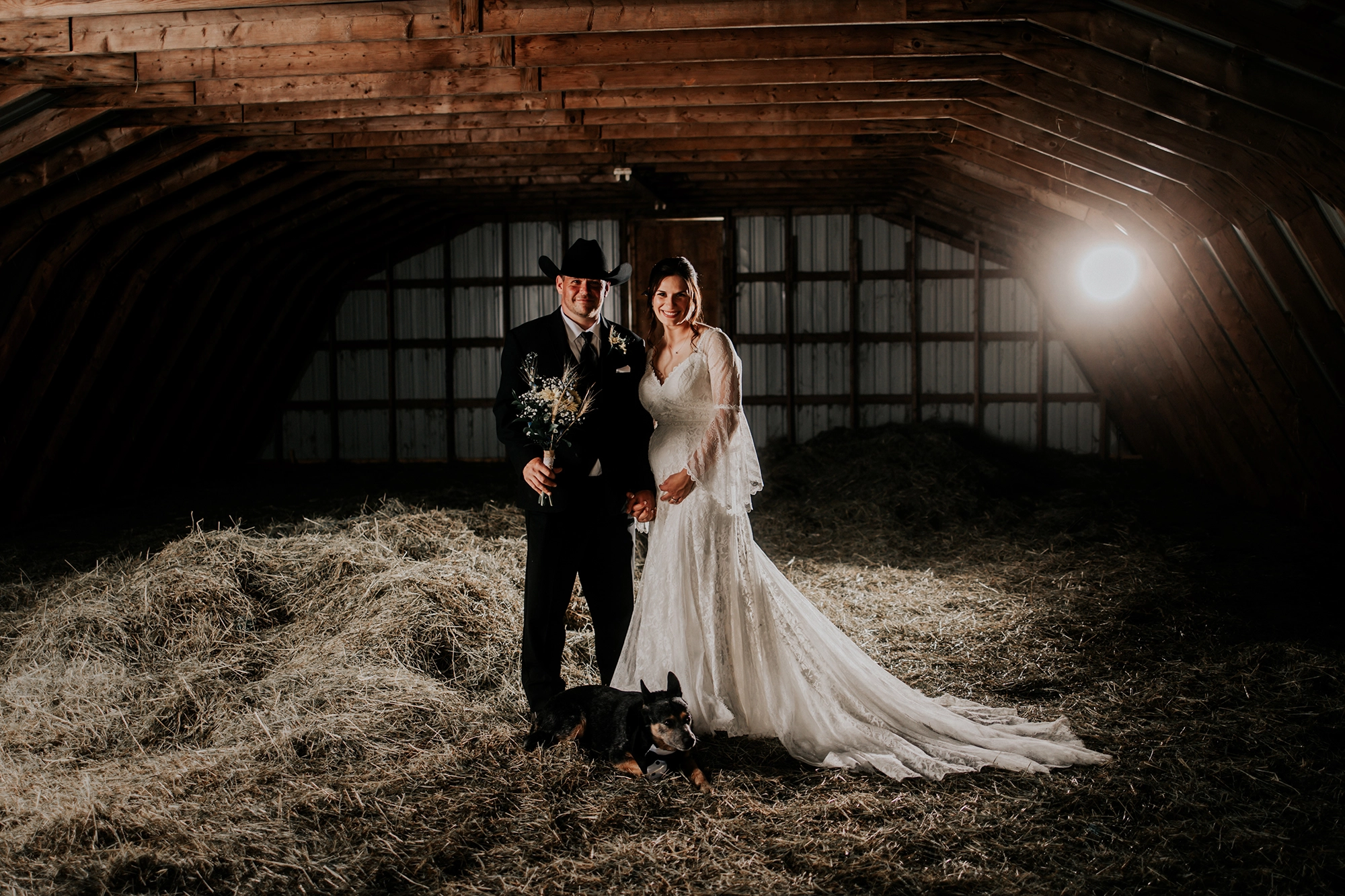 A moody, cinematic portrait of the couple in a rustic barn setting, captured in a dark fine art style.