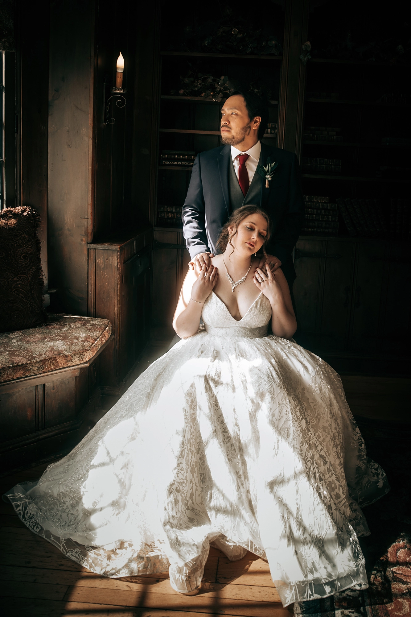 A moody, fine art wedding portrait of the bride seated with closed eyes as the groom gently places his hand on her shoulder, bathed in soft window light.