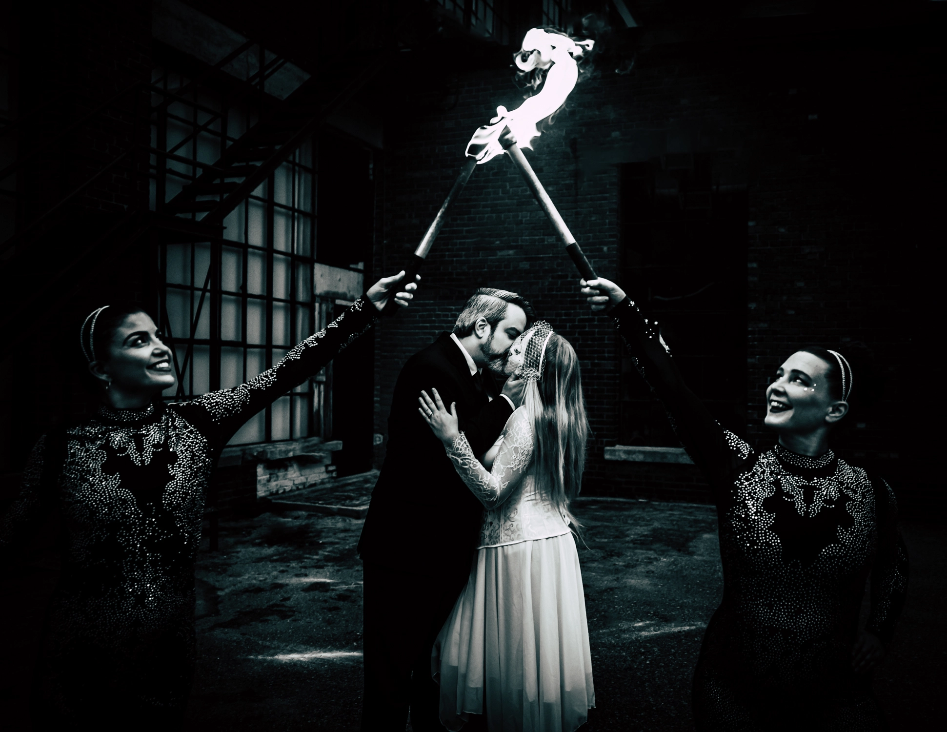 A dramatic black and white wedding photo of a couple kissing behind two fiery torches held by fire dancers.