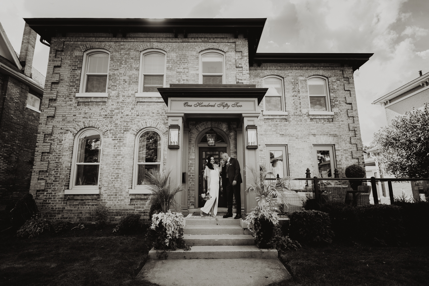 A timeless black and white portrait of a wedding couple in front of Sarah St. Amand’s elegant Brantford design house during their micro wedding.