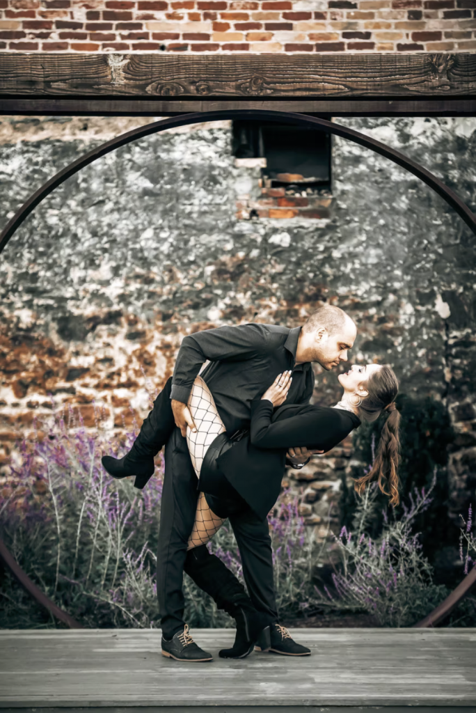 Couple wearing all black infront of rustic wall doing deep and about to kiss.